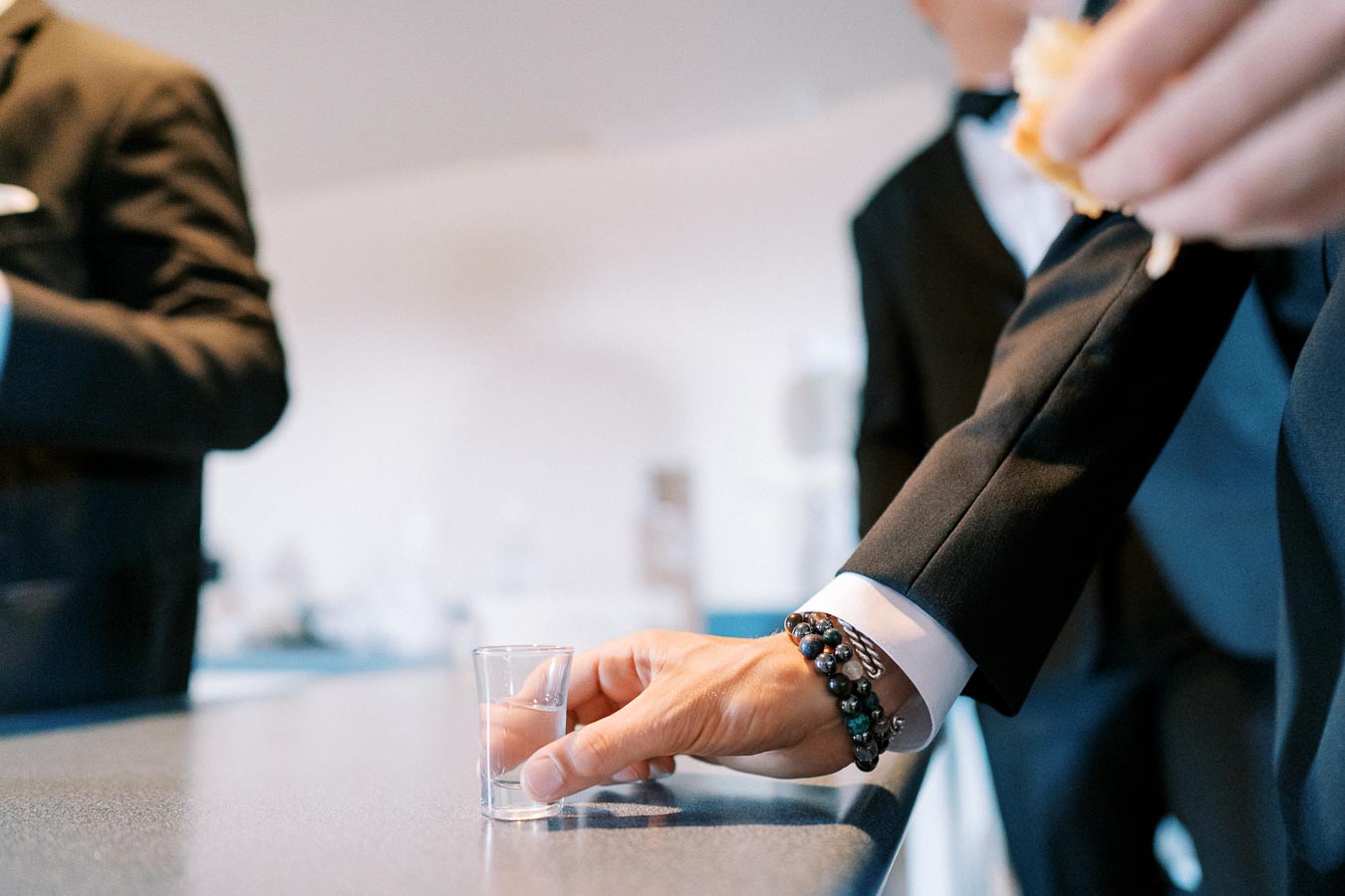 A person in formal attire reaching for a shot glass on a counter, wearing a stylish bracelet.