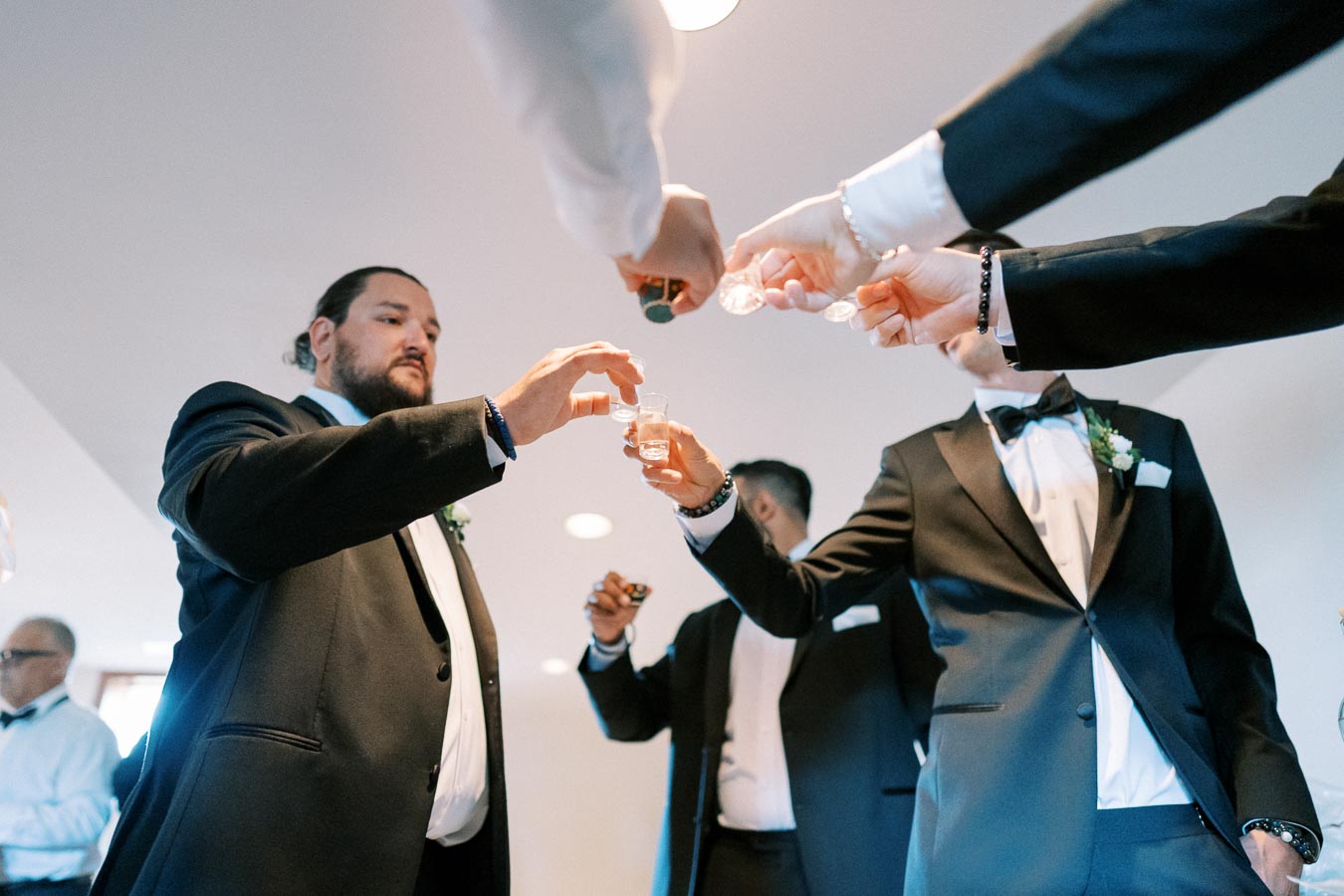 Groom and groomsmen in tuxedos raising glasses for a toast, capturing a celebratory moment before a wedding ceremony.