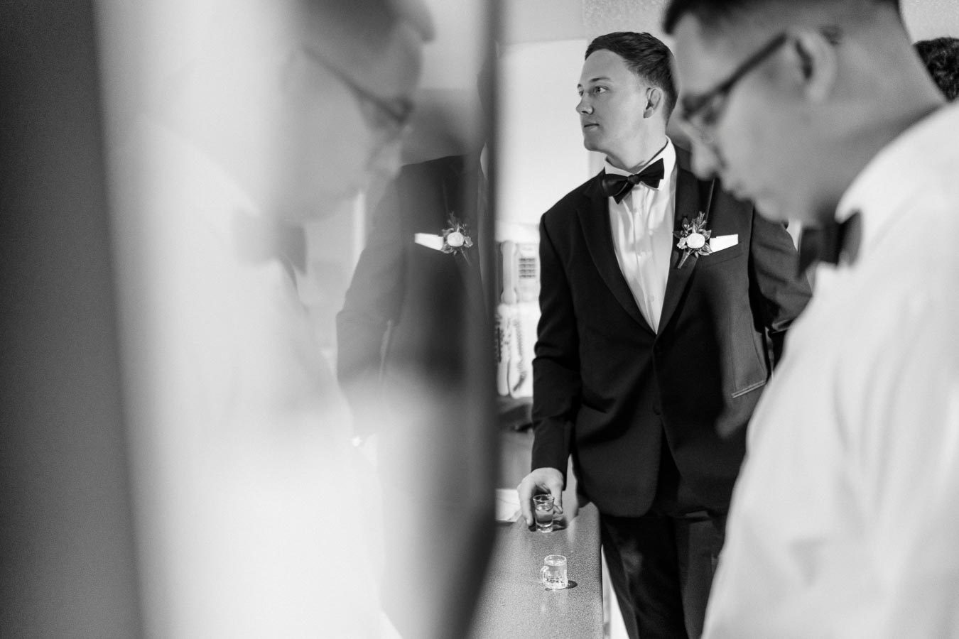 Black and white image of a groom preparing nervously in a tuxedo, holding a shot glass, with friends in the background before a wedding ceremony.