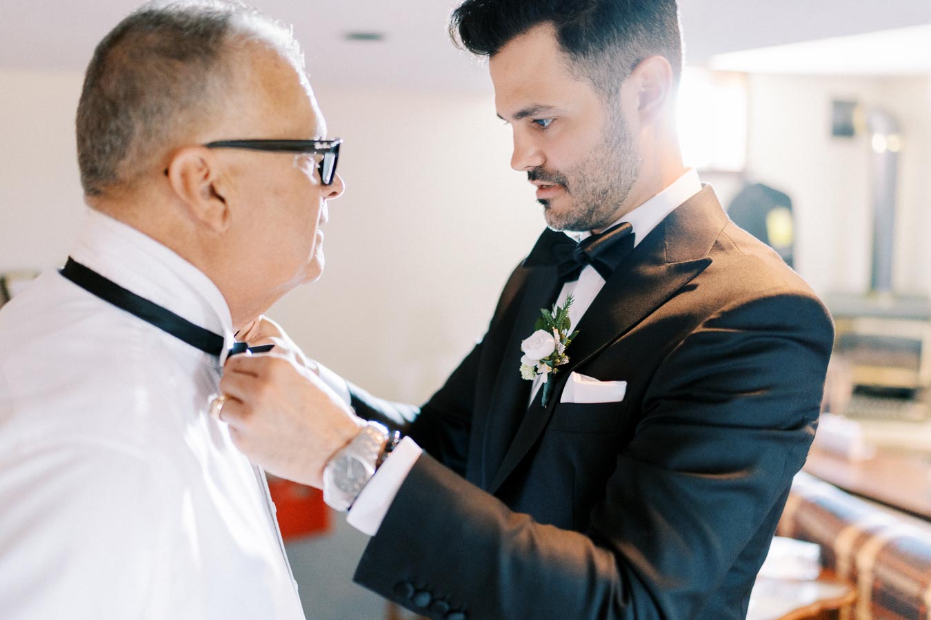A groom in a black tuxedo adjusts an older man's bow tie in a warmly lit room, highlighting a tender moment before a wedding ceremony.