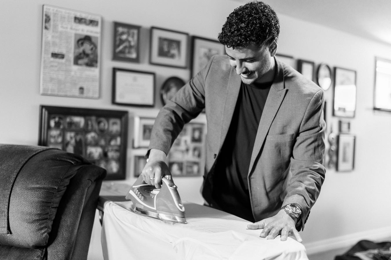 A man in a suit happily ironing a shirt at home, with a gallery wall of framed pictures in the background.