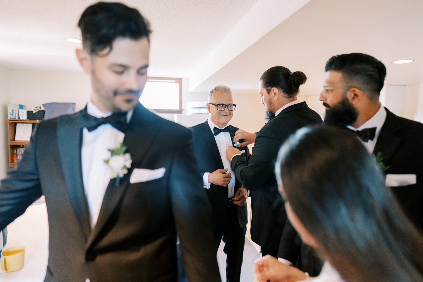 Group of groomsmen in tuxedos preparing for a wedding, adjusting each other's bow ties and boutonnieres in a bright room.