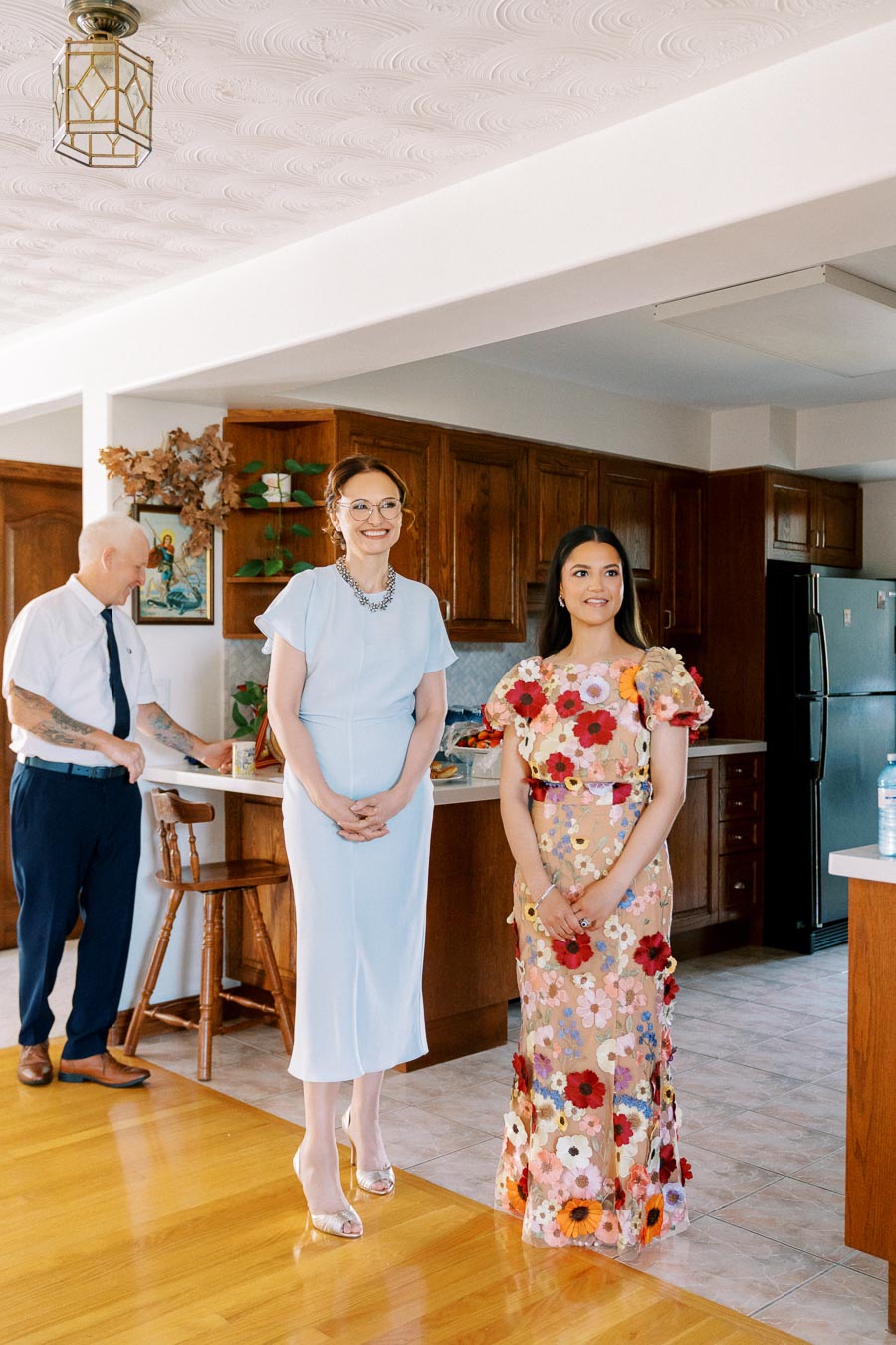 A cheerful group of people gathered in a warmly lit kitchen, featuring two women standing in the foreground wearing elegant dresses, one in light blue and the other in a colorful floral-patterned dress, while a man in a white shirt and tie is in the background near a wooden counter.