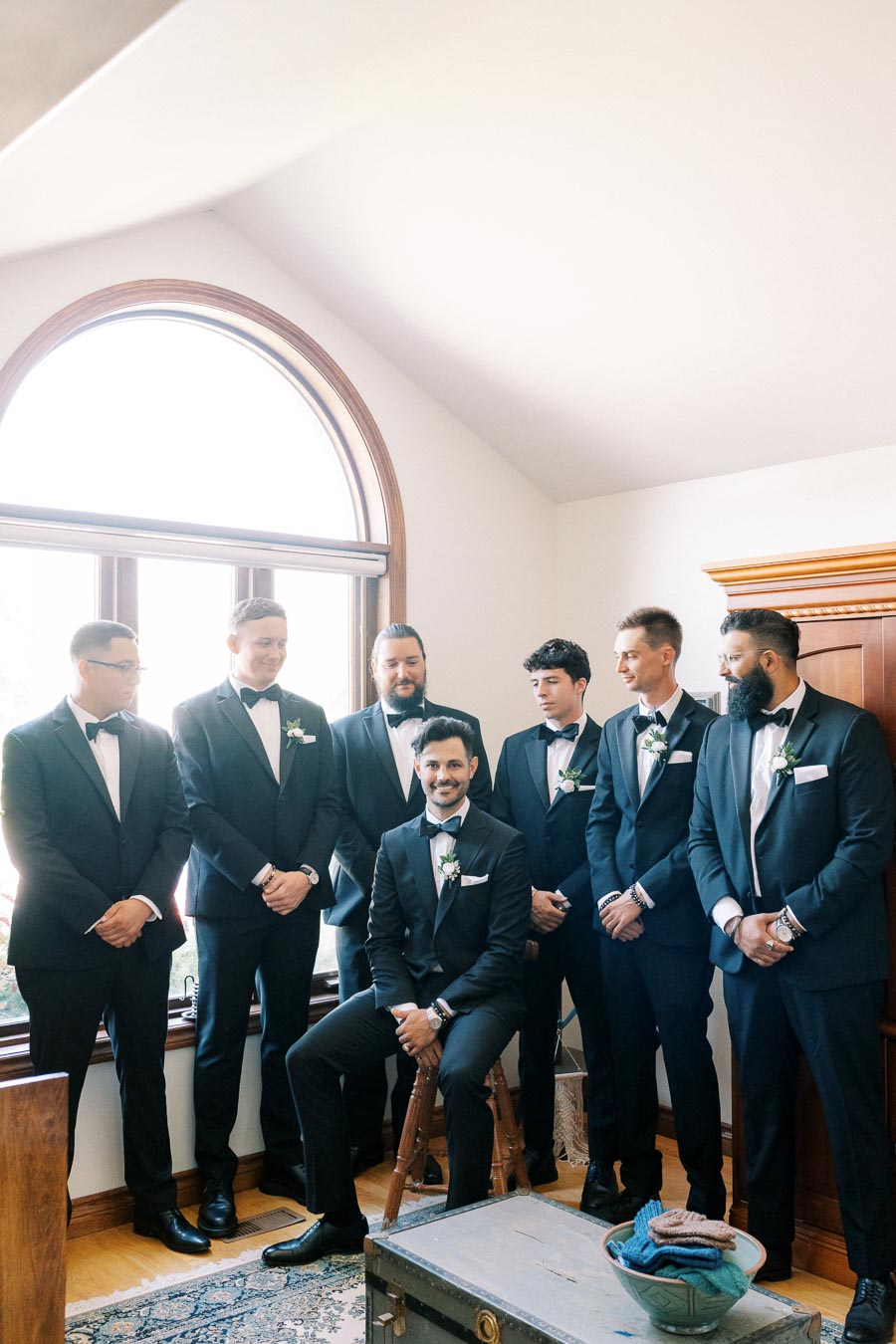 Groom with groomsmen in navy tuxedos posing indoors before a wedding ceremony, standing and sitting near a large arched window with natural light illuminating the room.