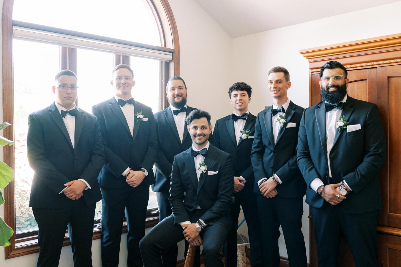 A group of seven groomsmen in matching black tuxedos and bow ties, standing indoors by a large window, posing for a wedding photo.