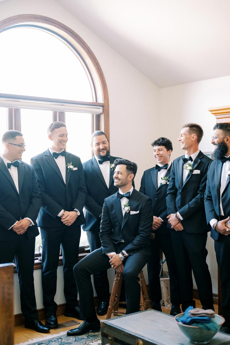 Group of groomsmen in black tuxedos smiling indoors, gathered around the seated groom near a large arched window.