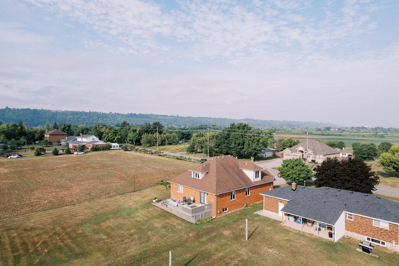 Aerial view of suburban neighborhood with brick houses surrounded by open fields and greenery under a partially cloudy sky.