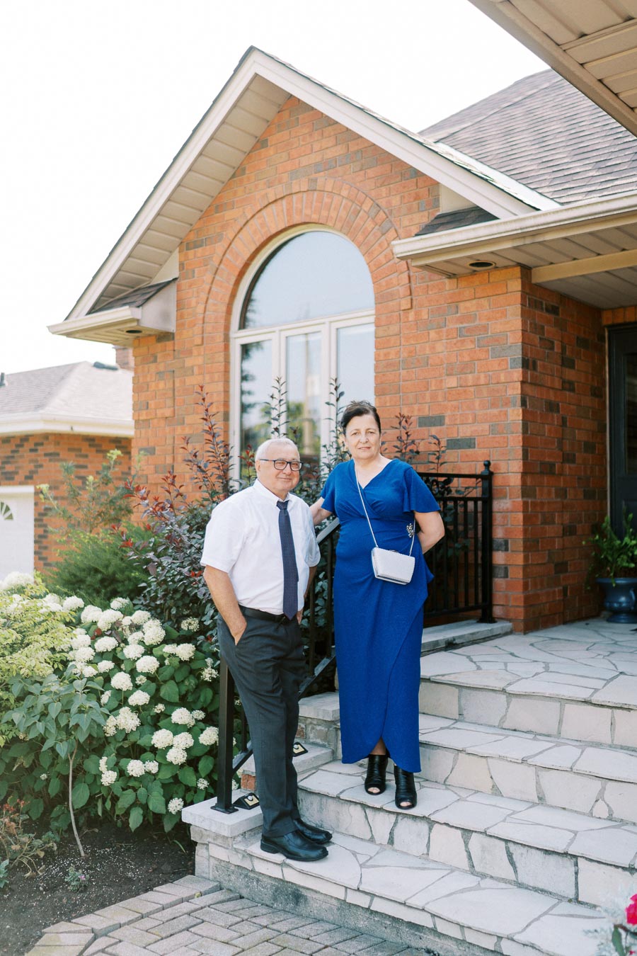 Senior couple standing on the front steps of a brick house with a garden in the background.