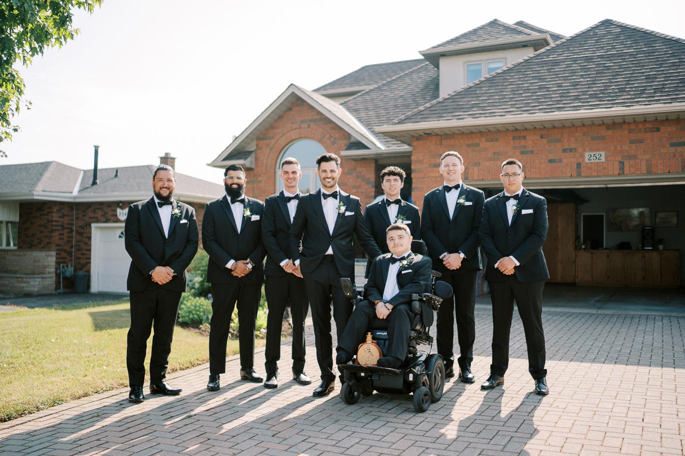 A group of groomsmen in black tuxedos standing on a driveway, with one seated in a wheelchair, posing in front of a brick house on a sunny day.