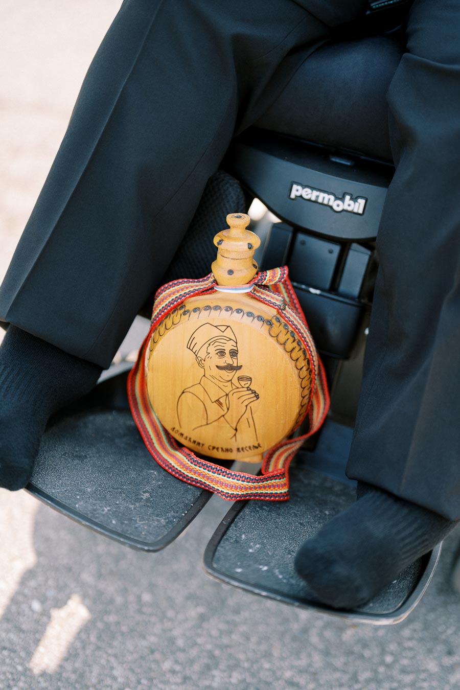 A wooden flask with an engraved drawing of a man holding a glass, wrapped in a colorful striped fabric, resting on a wheelchair footrest.