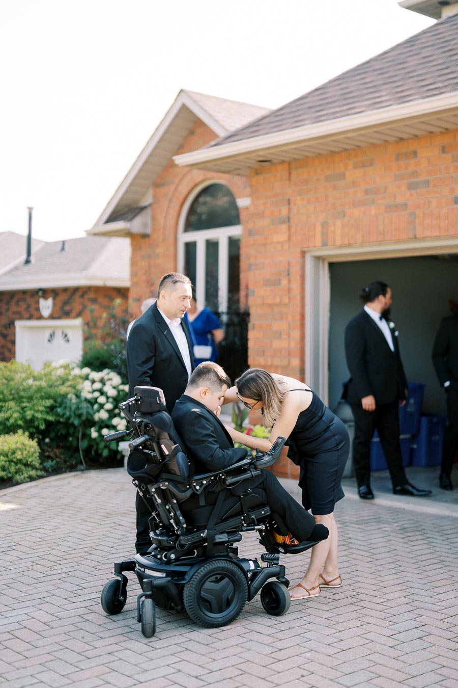 A group of people gathered outside a brick house, with a woman adjusting the suit of a man in a wheelchair, and others nearby dressed formally.