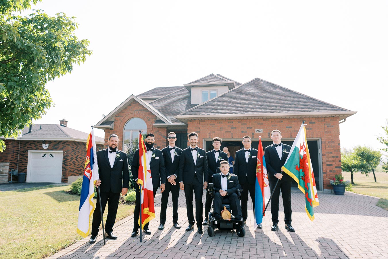 Groomsmen in formal suits holding various national flags, standing in front of a brick house, outdoor wedding photo.