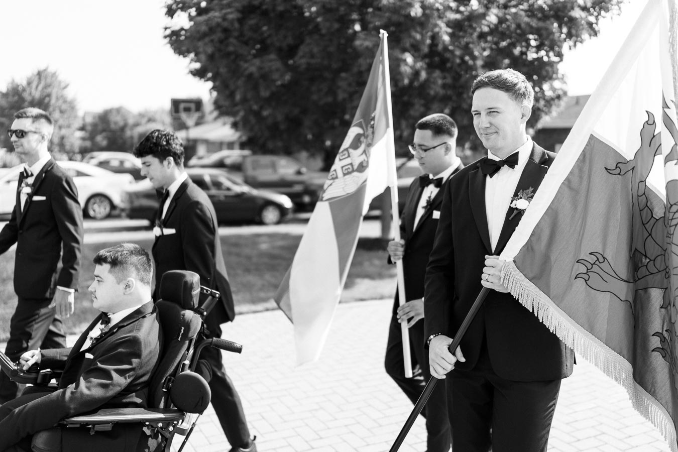 A group of men in formal attire walking outdoors, two of them holding flags, one in a wheelchair, under a sunny sky with trees and cars in the background. Black and white photograph.