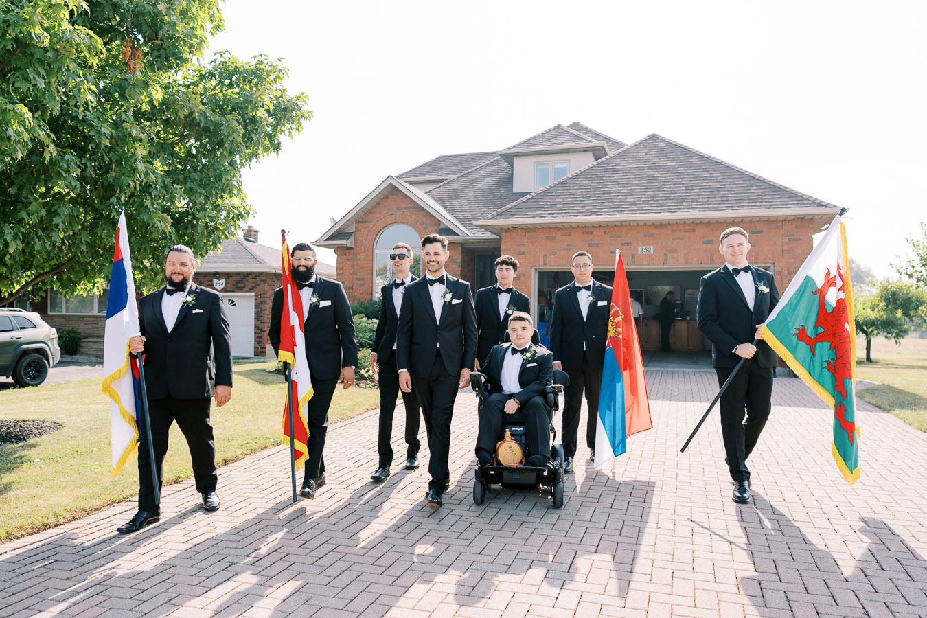 A group of groomsmen in formal suits standing on a brick driveway, holding various national flags, in front of a suburban house on a sunny day.