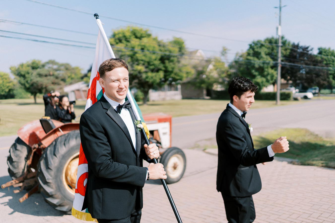 Two men in formal suits participate in an outdoor event, one holding a flag, with a red tractor in the background on a sunny day.
