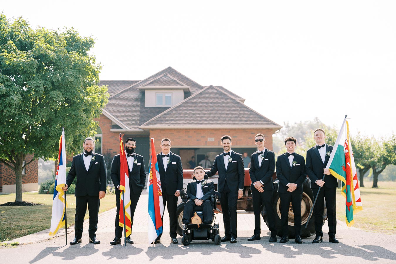 A group of groomsmen in formal tuxedos, standing alongside a person in a wheelchair, holding various national flags, in front of a brick house.