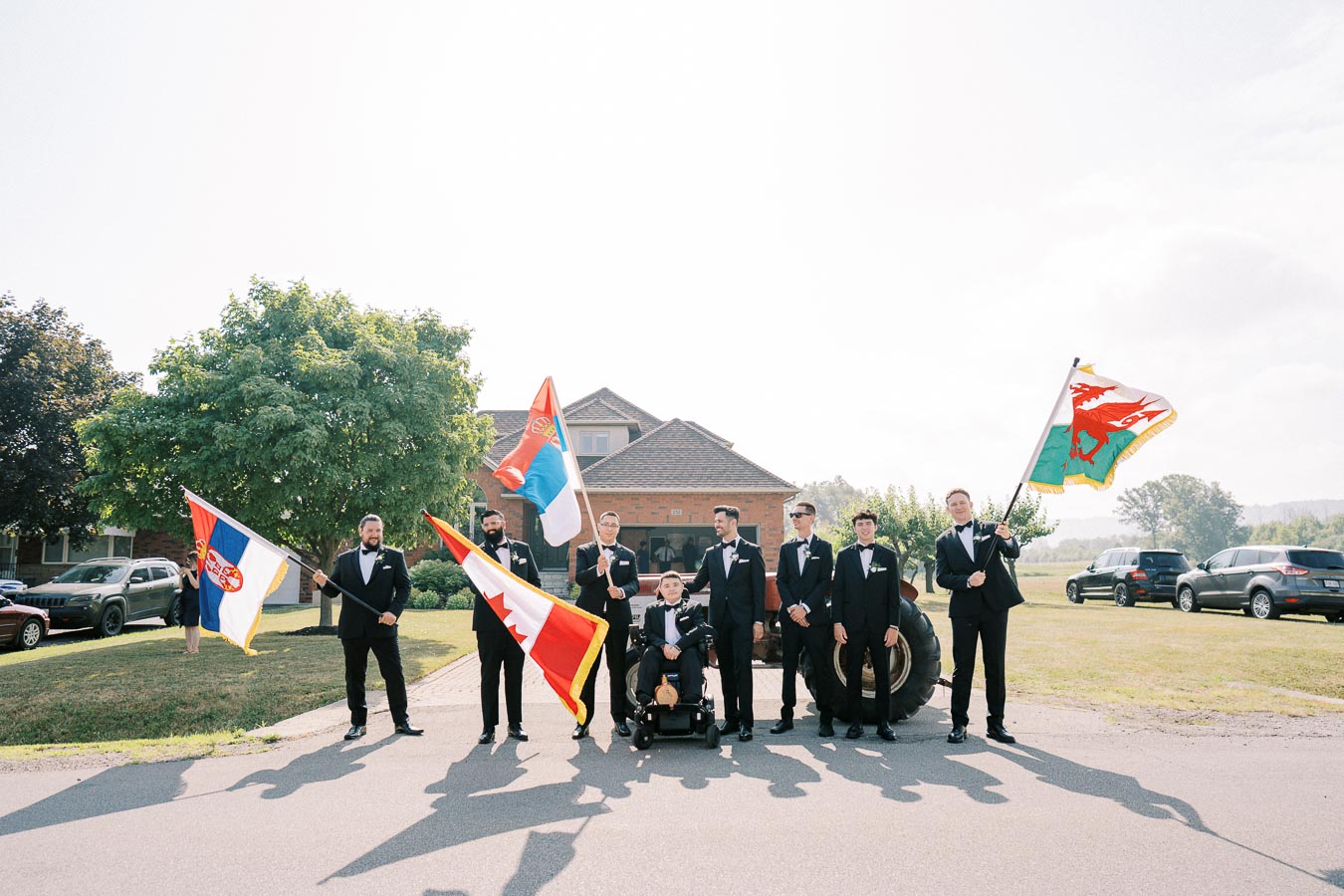 A group of men in formal attire poses outdoors, holding various international flags, with a brick house, trees, and cars in the background on a sunny day.