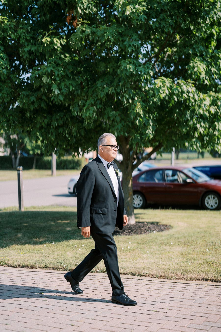 An older gentleman in a black suit and bow tie walking on a sunny day, surrounded by green trees and a parked car in the background.
