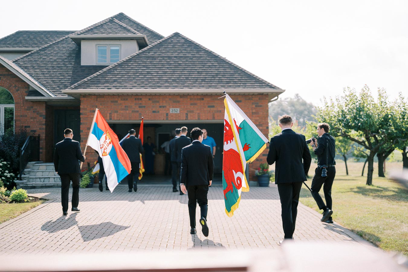 A group of people in formal attire walking towards a house, carrying flags from different countries, on a sunny day.