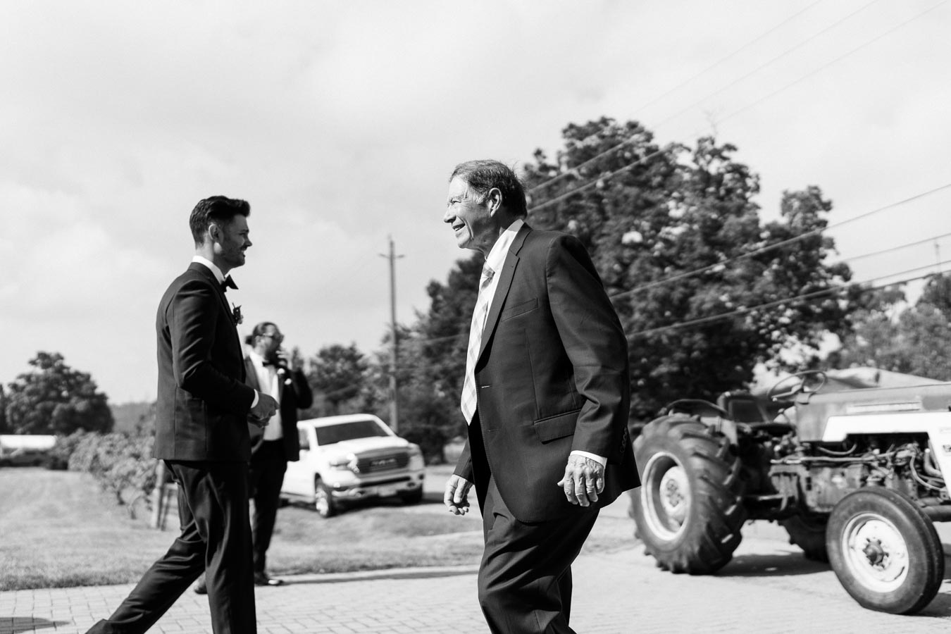 Black and white image of two men in suits smiling and walking outdoors, with a vintage tractor and parked vehicle in the background.