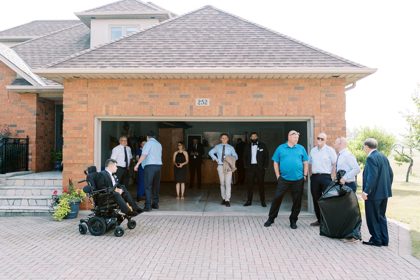 A group of people gathered outside a brick house with an open garage, featuring a person in a wheelchair, during a social event on a sunny day.