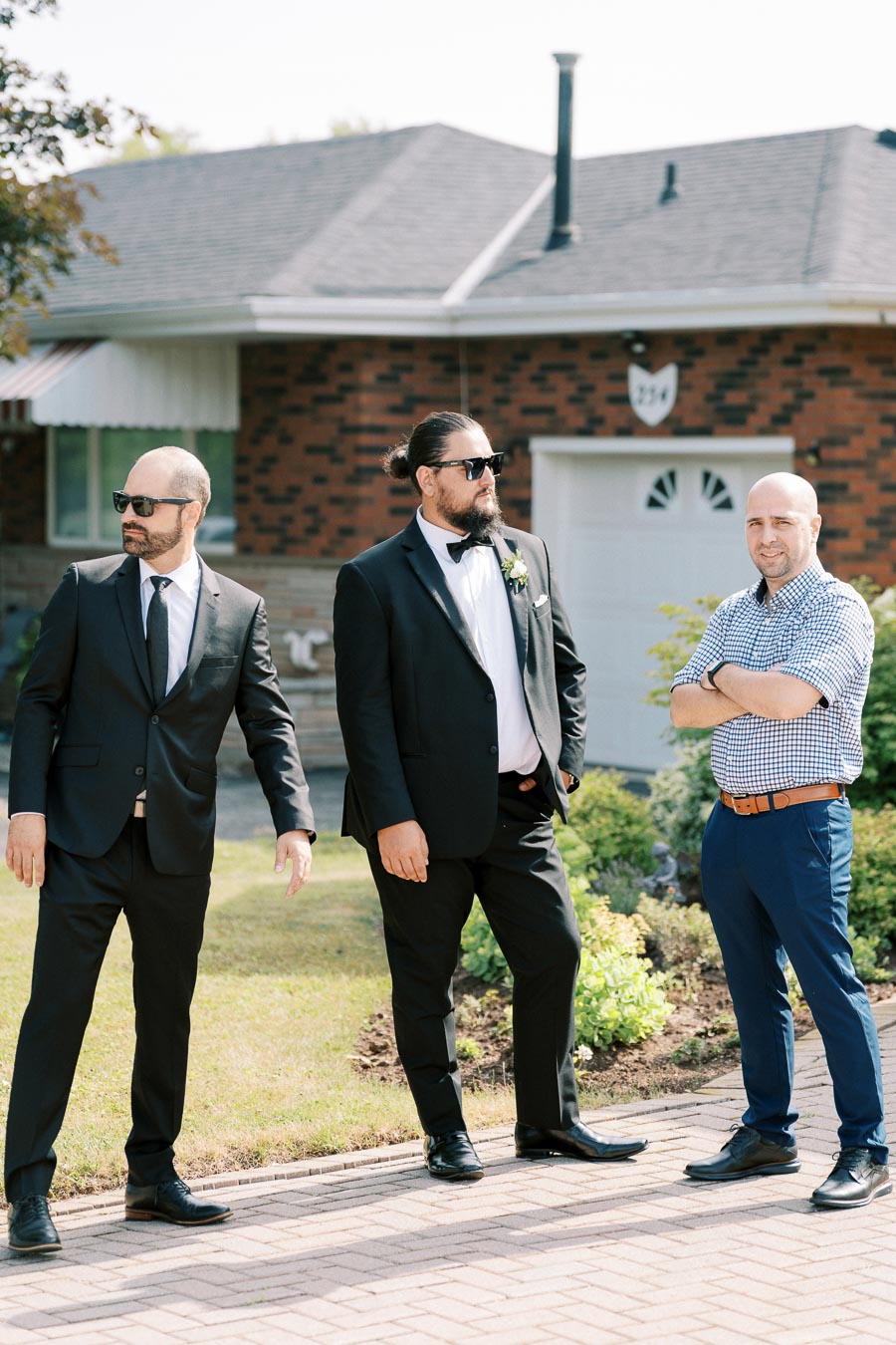 Three men, two in formal black suits and one in a checkered shirt, stand outdoors on a sunny day in front of a brick house.