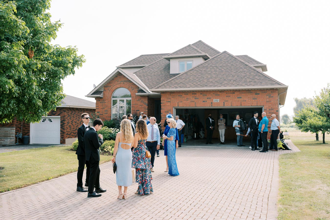 Group of people gathered outside a brick house, engaging in conversation during a social event on a sunny day, with a well-kept lawn and trees in the background.
