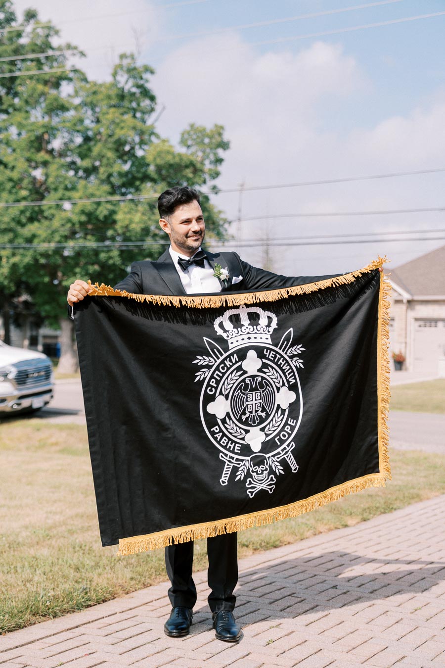 A man in a suit holds a large black flag with white designs and text, standing on a sunny outdoor sidewalk, with trees and houses in the background.