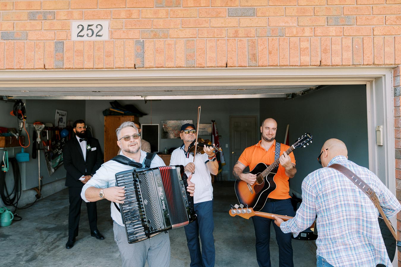 A group of musicians playing instruments, including an accordion, violin, acoustic guitar, and bass, in a residential garage setting. The lively scene captures a casual neighborhood gathering with instruments and engaged individuals.