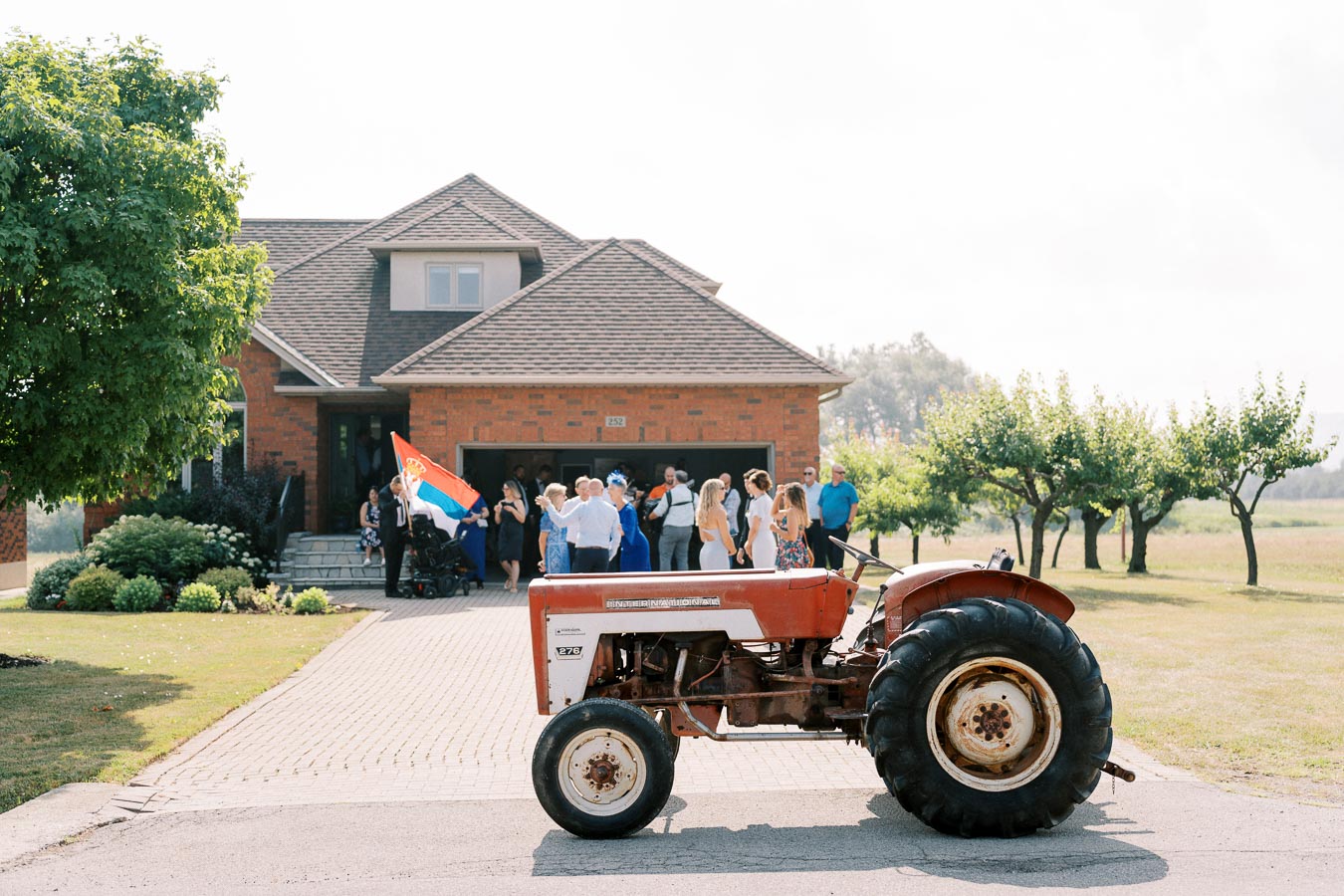 A red tractor parked on a driveway in front of a red brick house, with a group of people gathered outside. One person is holding a flag, and there are trees and a lawn in the background on a sunny day.