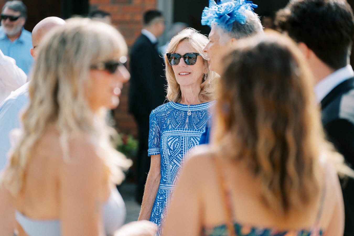 Elegant woman in a blue patterned dress and sunglasses at an outdoor event, surrounded by people in stylish attire.