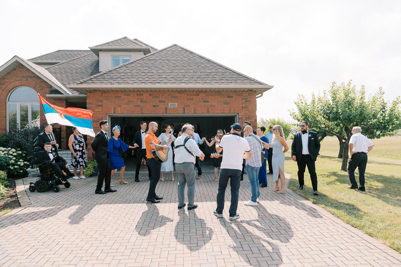 A group of people celebrating outside a red brick house, with one person holding a Serbian flag, while others play musical instruments and dance on a sunny day.
