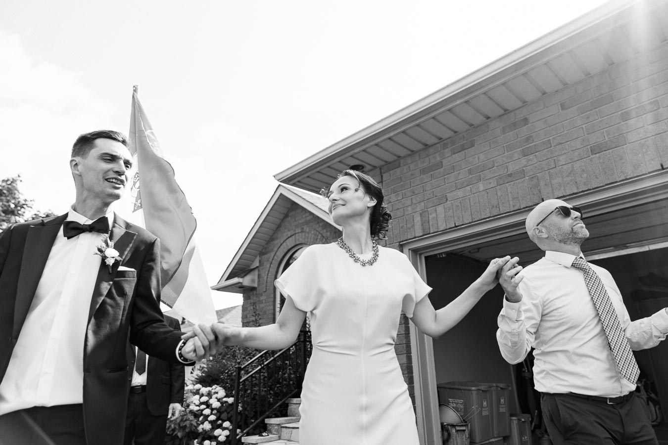 A black and white photo of three elegantly dressed people holding hands outside a brick house, portraying joy and celebration, with a focus on a woman in a dress and a man in a suit with a boutonniere.