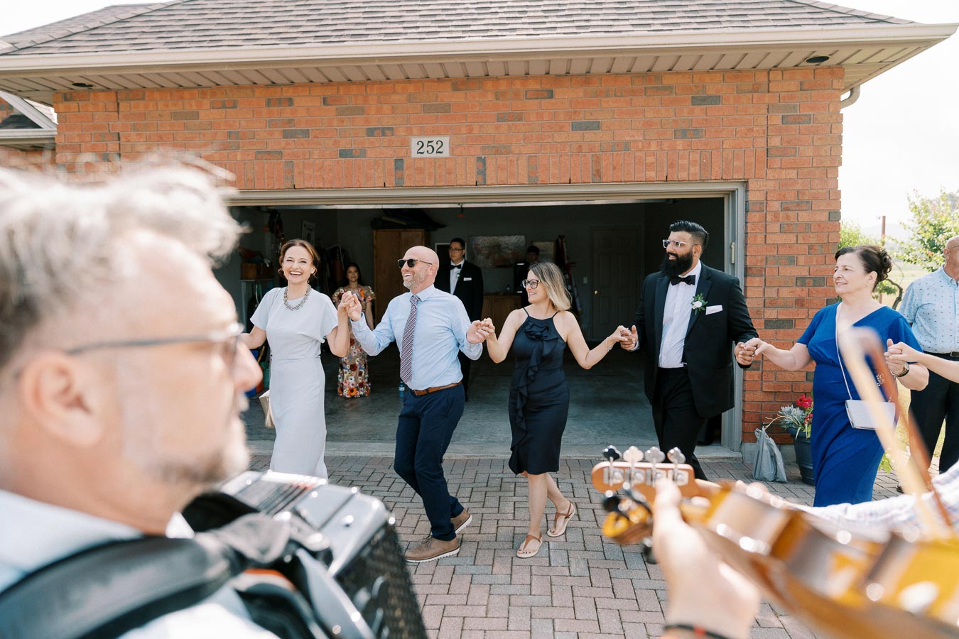 A group of people joyfully dancing hand-in-hand outdoors near a brick house, with musicians playing instruments in the foreground, creating a festive atmosphere.