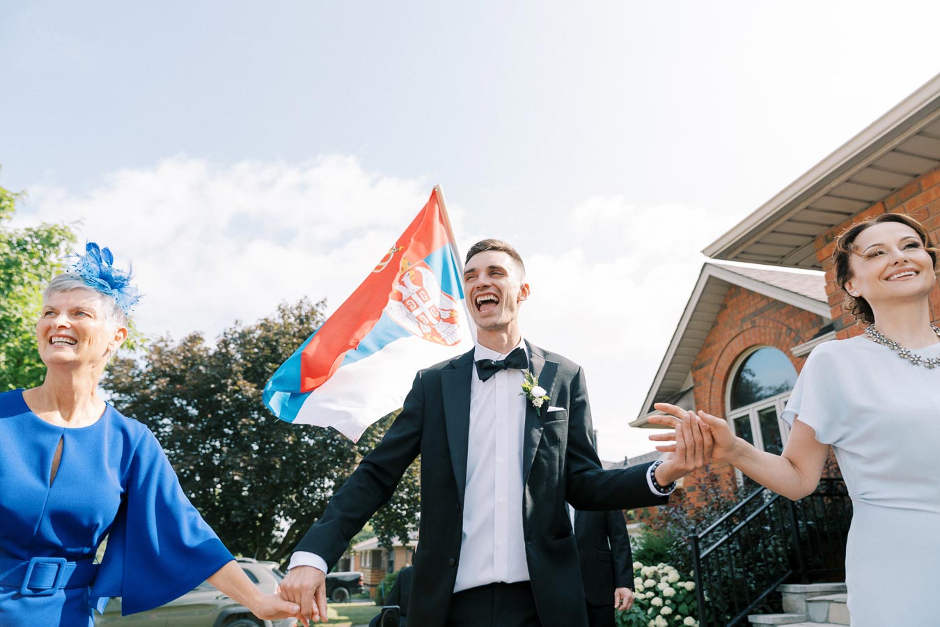 Smiling groom in a black tuxedo holding hands with two women at a wedding celebration, in front of a house with a Serbian flag flying behind.
