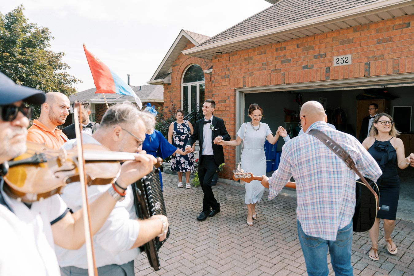 Outdoor celebration with people dancing in a circle, accompanied by musicians playing violin and guitar, in front of a brick house.