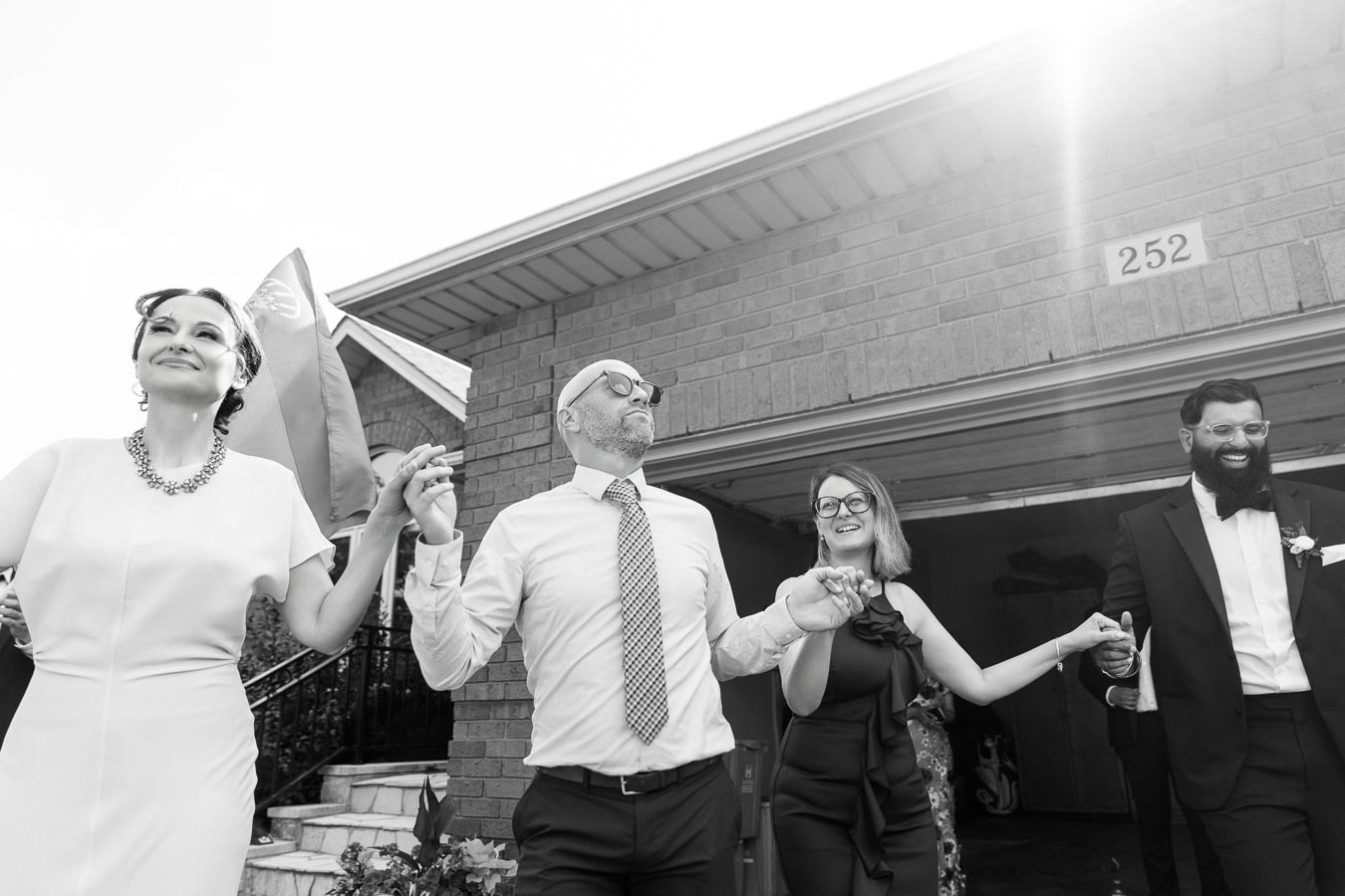 A group of people joyfully holding hands outside a house, with one person holding a flag, dressed in formal attire. Bright sunlight enhances the black and white image, creating a lively atmosphere.