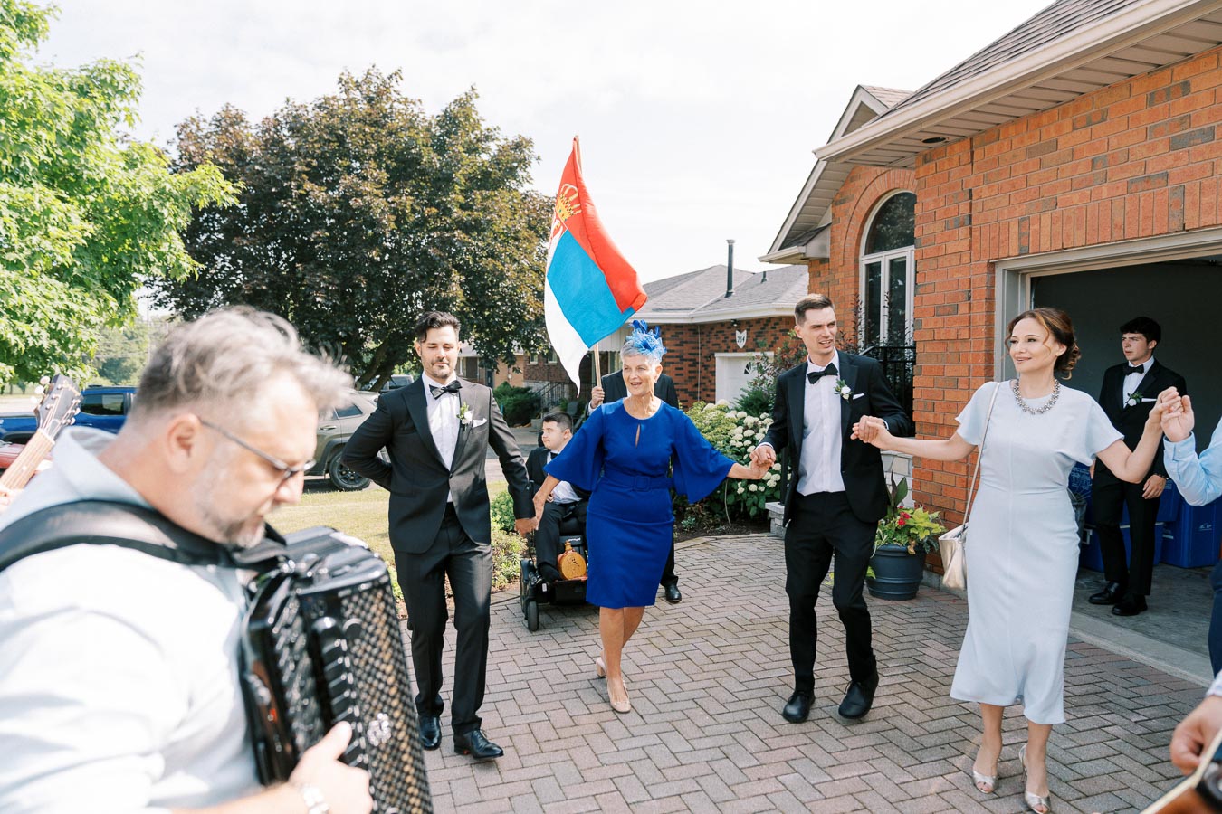 A joyful outdoor celebration with people dressed elegantly in formal attire, holding hands in a dance. A musician plays the accordion in the foreground, while a person holds a flag in the background, set against a brick house and lush greenery.