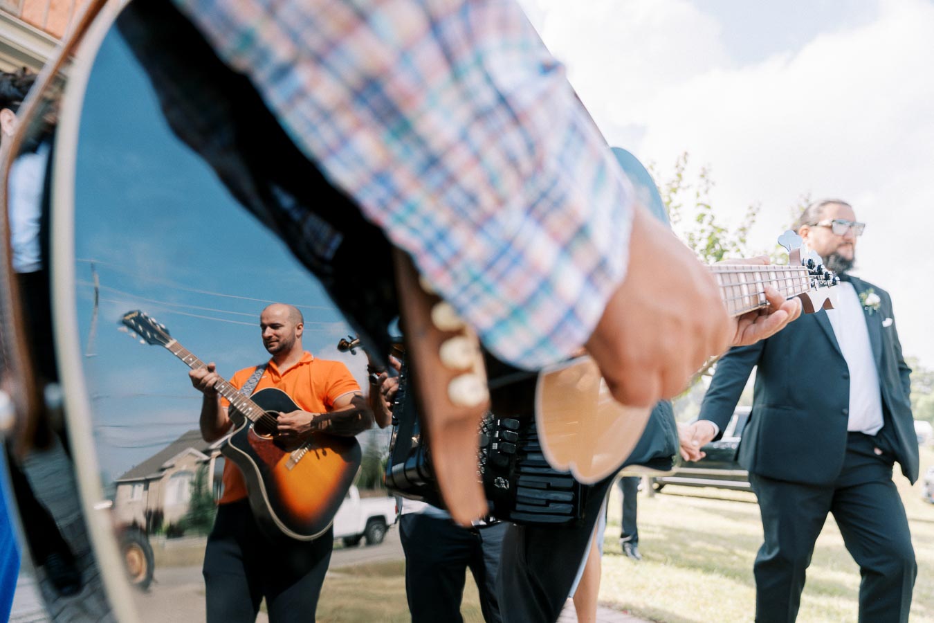 A vibrant outdoor scene featuring a close-up of a musician playing a string instrument, with another musician reflected on the guitar's surface. In the background, a person in formal attire, possibly at a wedding or event, under a clear blue sky.