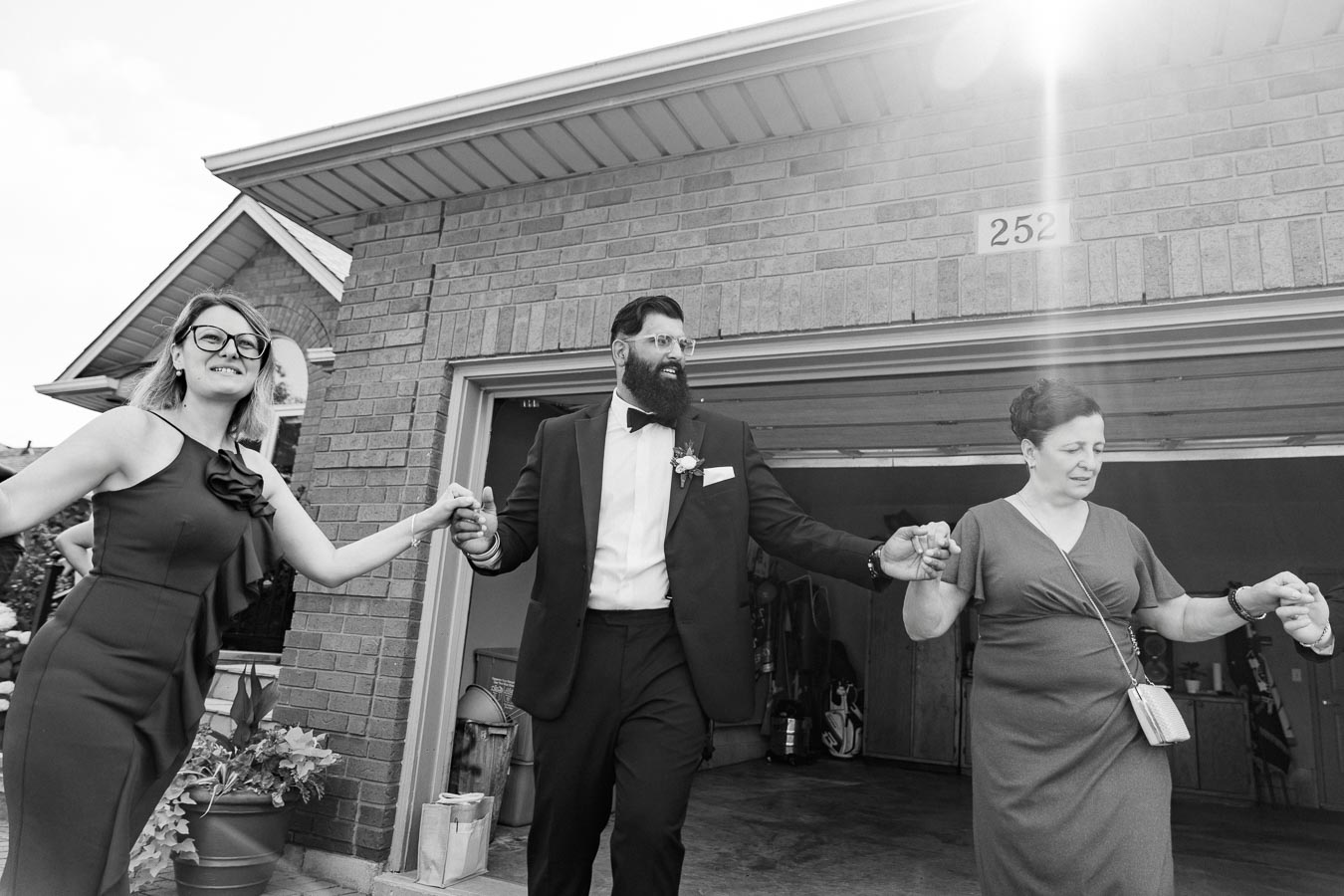 A black and white photo of three people holding hands in front of a brick house, with a man in a suit and two women in dresses, reflecting a joyful moment.
