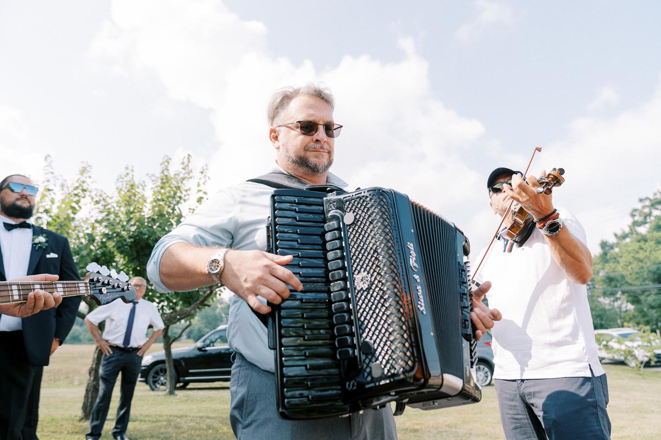 A group of musicians playing instruments outdoors, including an accordion, guitar, and violin, on a clear day with a backdrop of trees and a car.