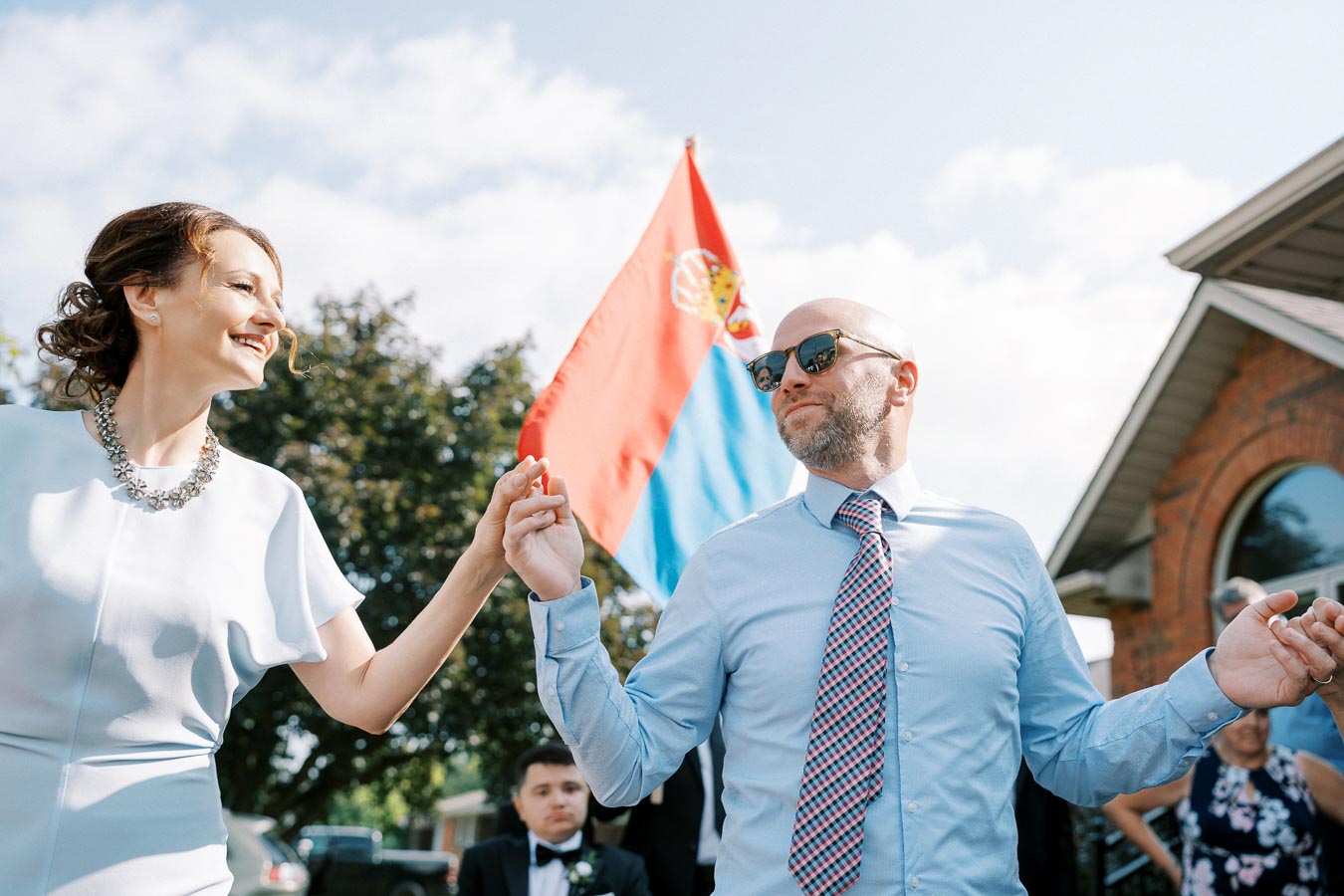 A couple dancing joyfully at an outdoor event, with the Serbian flag waving in the background, under a clear blue sky.