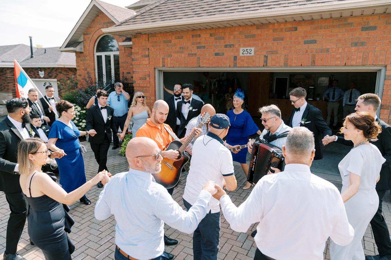 A group of people celebrating outdoors in a lively circle dance, featuring musicians playing guitar and accordion, in front of a brick house with a garage.