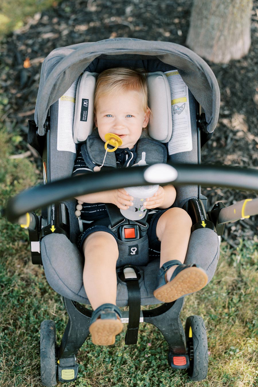 A smiling baby with a pacifier sits in a gray stroller outdoors, holding a bottle, enjoying a sunny day on the grass.