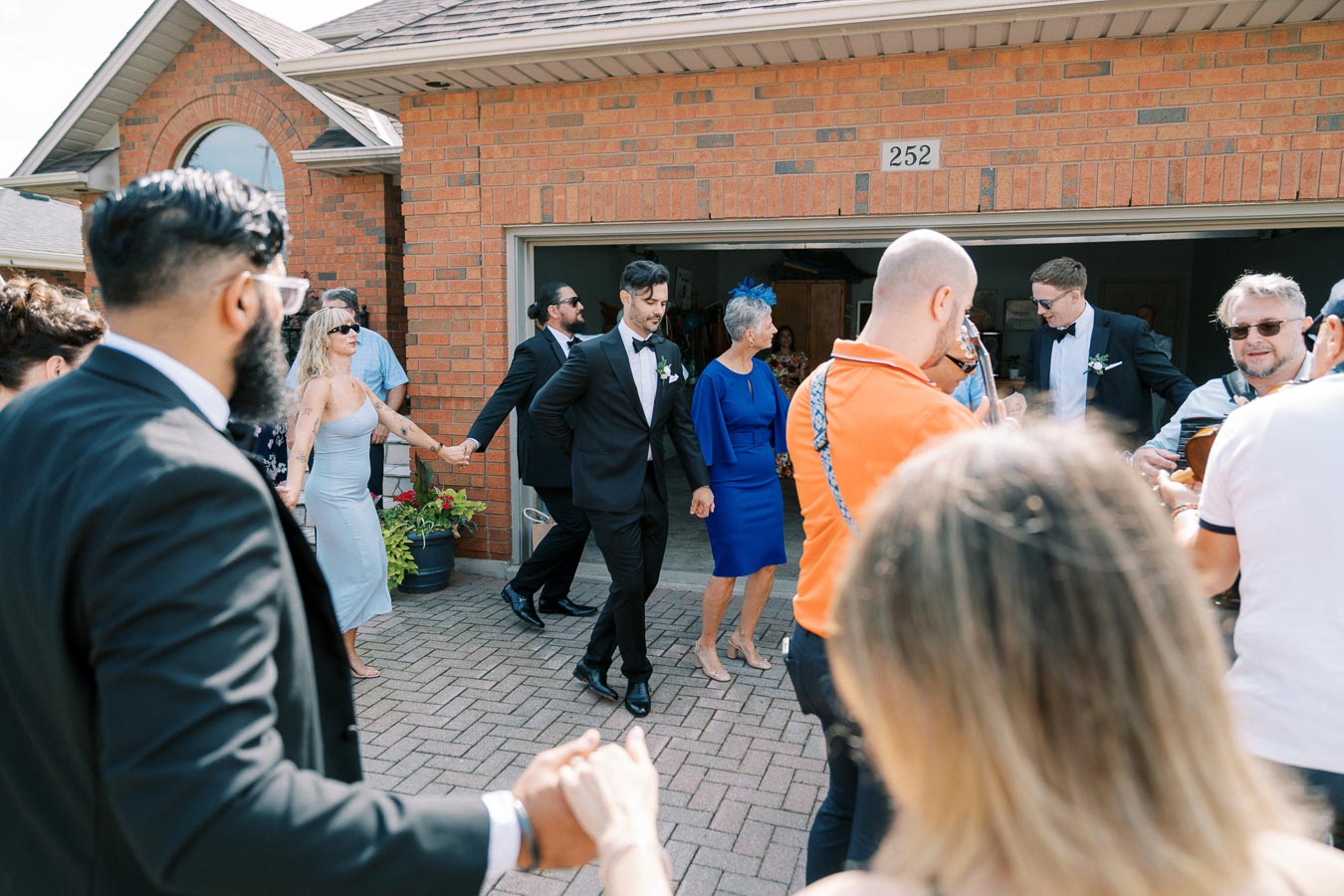 A wedding group dancing in a circle outside a brick house, featuring guests in formal attire enjoying a celebration under a sunny sky.