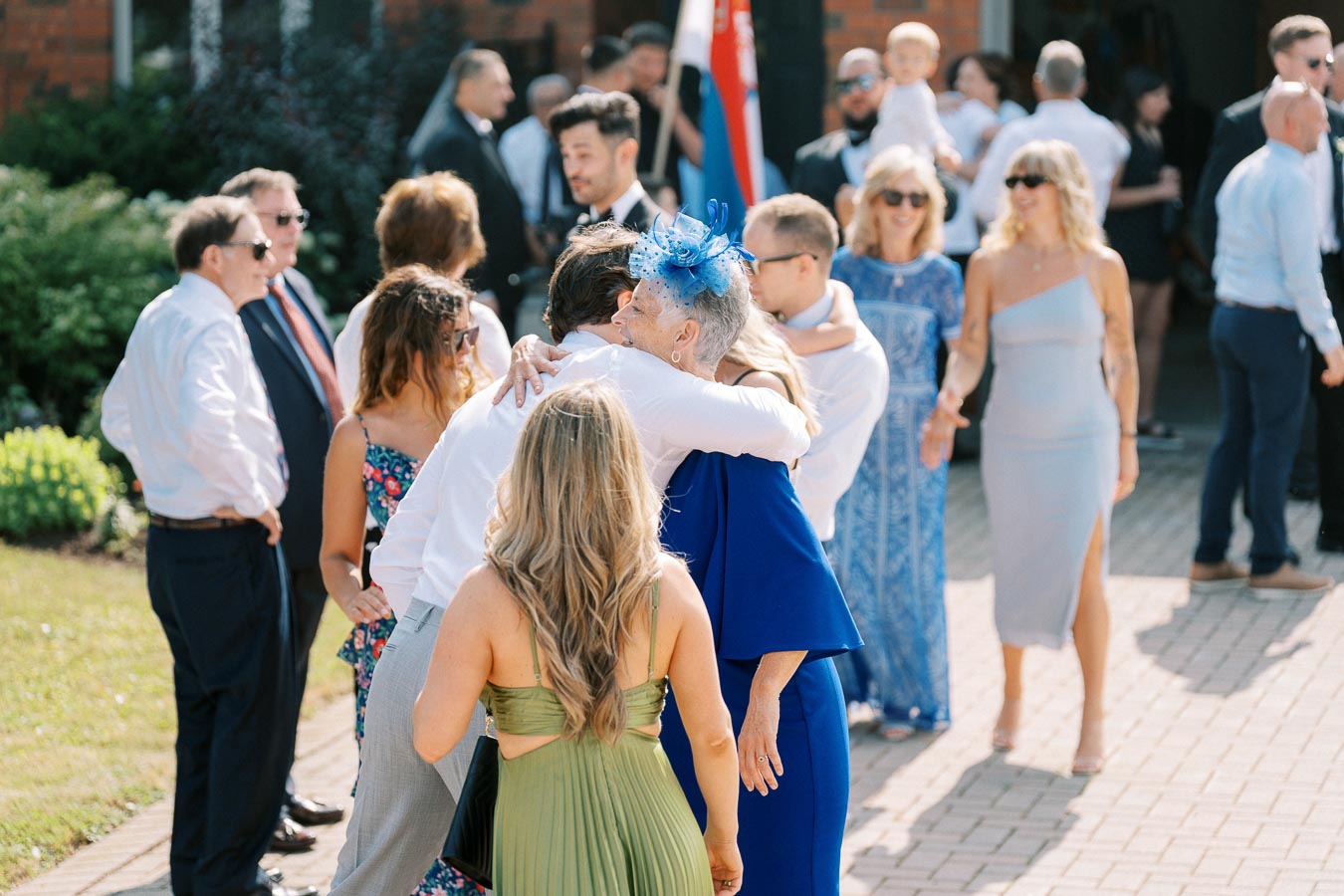 A group of elegantly dressed people socializing at an outdoor event, with a woman in a blue dress embracing a guest, surrounded by greenery and sunlight.