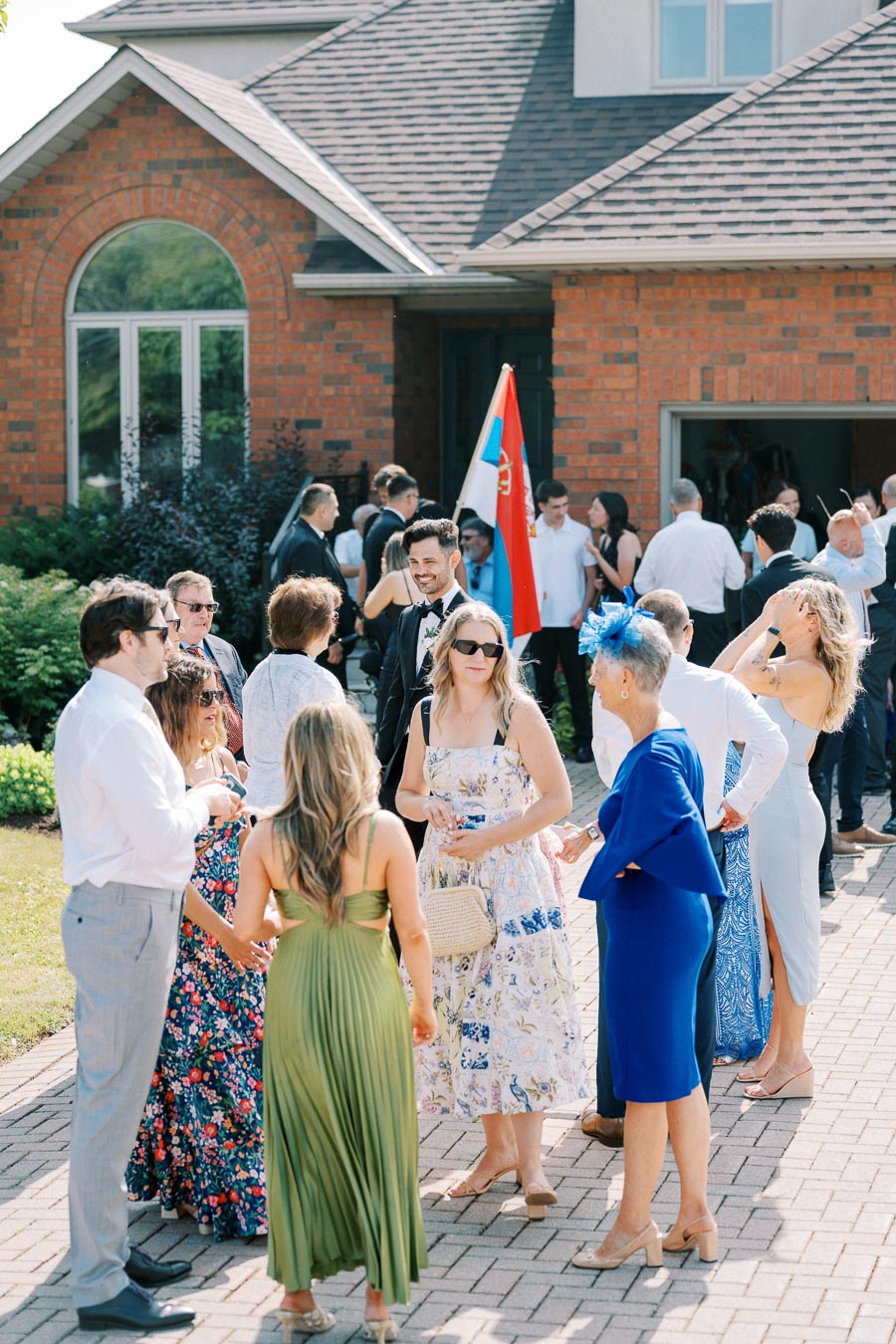 A group of elegantly dressed people socializing outdoors at a formal gathering, with a brick house in the background and a decorative flag visible, suggesting a special event or celebration.