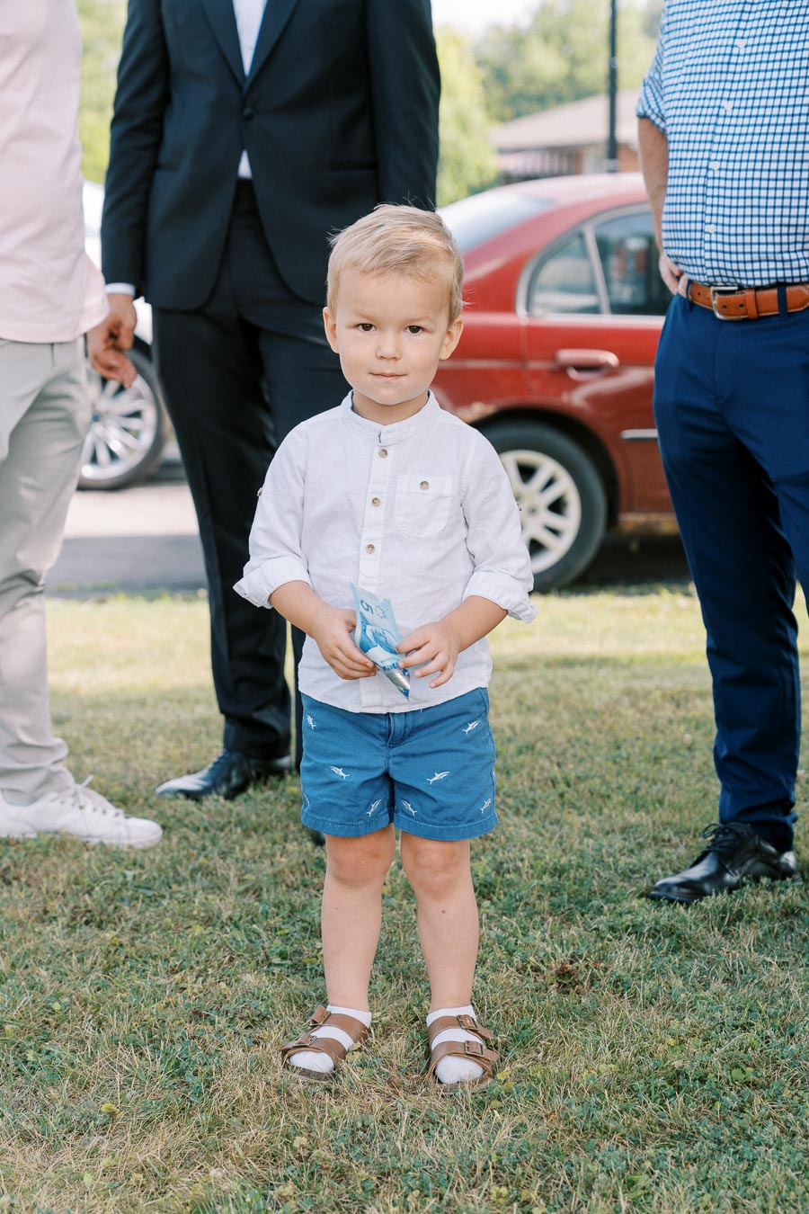 Young boy standing on grass holding play money, surrounded by adults in formal attire, with a red car and greenery in the background.