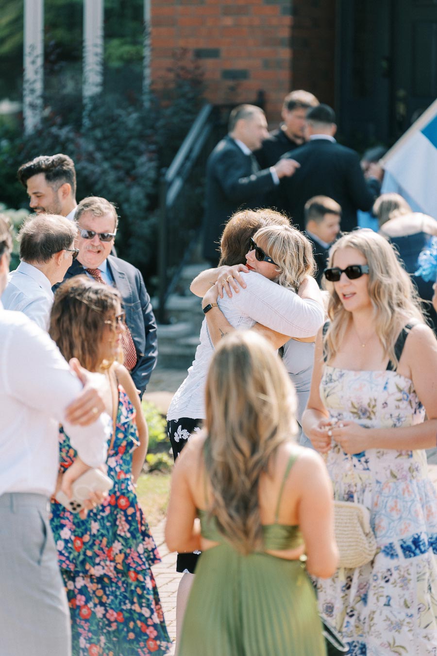 Group of people socializing and embracing outdoors during a sunny event, with individuals wearing summer attire and sunglasses.