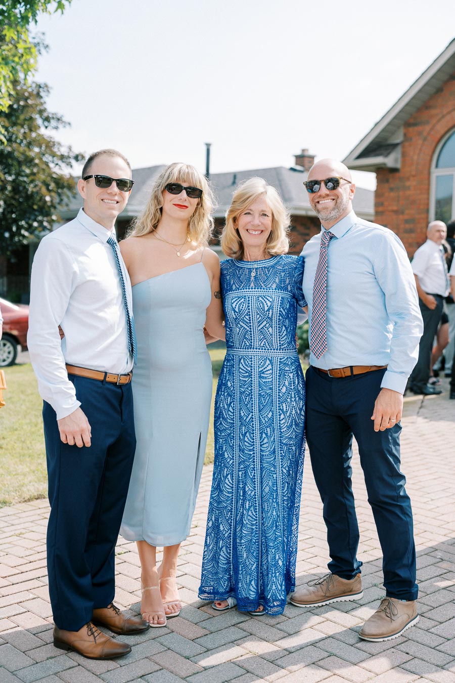 A group of four people dressed in formal attire, standing together outdoors on a sunny day. The women wear elegant blue dresses, while the men wear light shirts, ties, and sunglasses. A brick house and green lawn are visible in the background.
